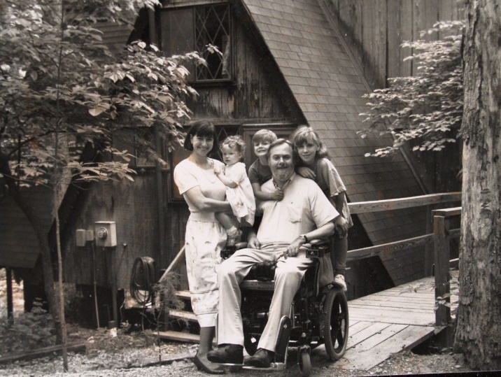 Black and white photo of the Starkloff family around 1990 smiling in front of a cabin. Colleen holds Emily as a toddler, while Maxim and Meaghan stand on the wheels of Max’s powerchair and lean on their father’s shoulders.
