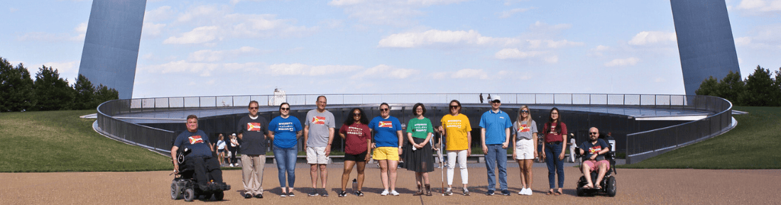 Community Education Page Banner Image – staff at Arch 2020 Starkloff staff and community members in colorful disability pride tshirts smiling in a line under the Gateway Arch