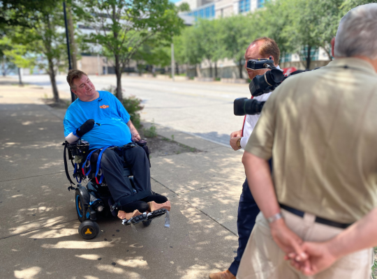 Steve KSDK Sidewalk Interview 2023 Steve seated in his power chair on a downtown sidewalk giving a TV news interview.