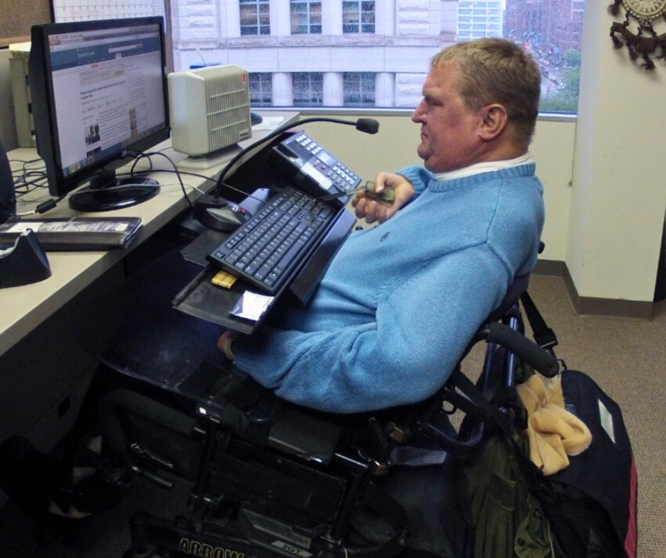 Steve, a power wheelchair user, works in his office. His desk is elevated and there is a microphone positioned directly at his mouth.