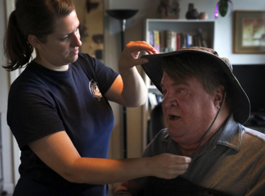 Steve getting ready for the day with PCA Steve seated in his power wheelchair, talking with his PCA who is assisting him in getting ready to head to work. She is adjusting his hat for sun protection.
