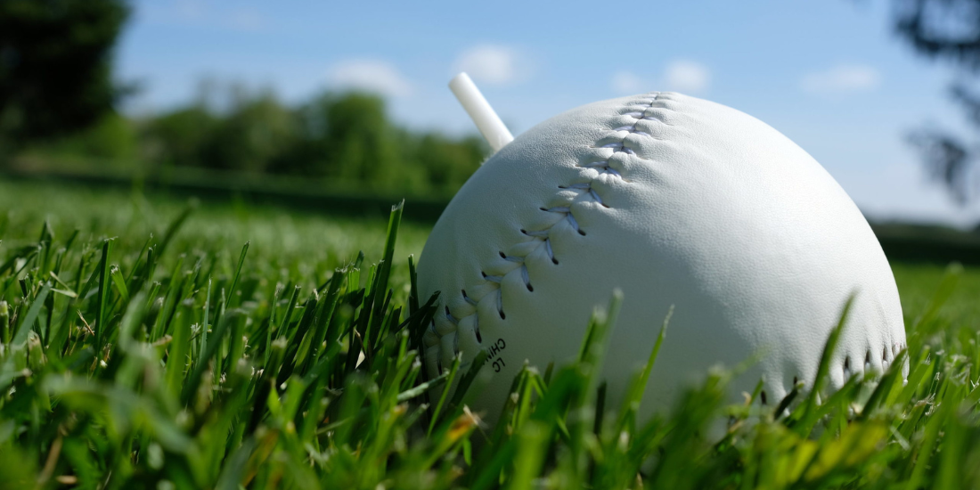 A beep baseball sitting in the grass. It is a bit larger than a softball and has a plastic post sticking out of the top.