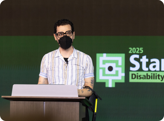 Dom Kelly delivering opening keynote. He is wearing a black N95 mask and has the loop of a crutch hanging from his visible elbow.