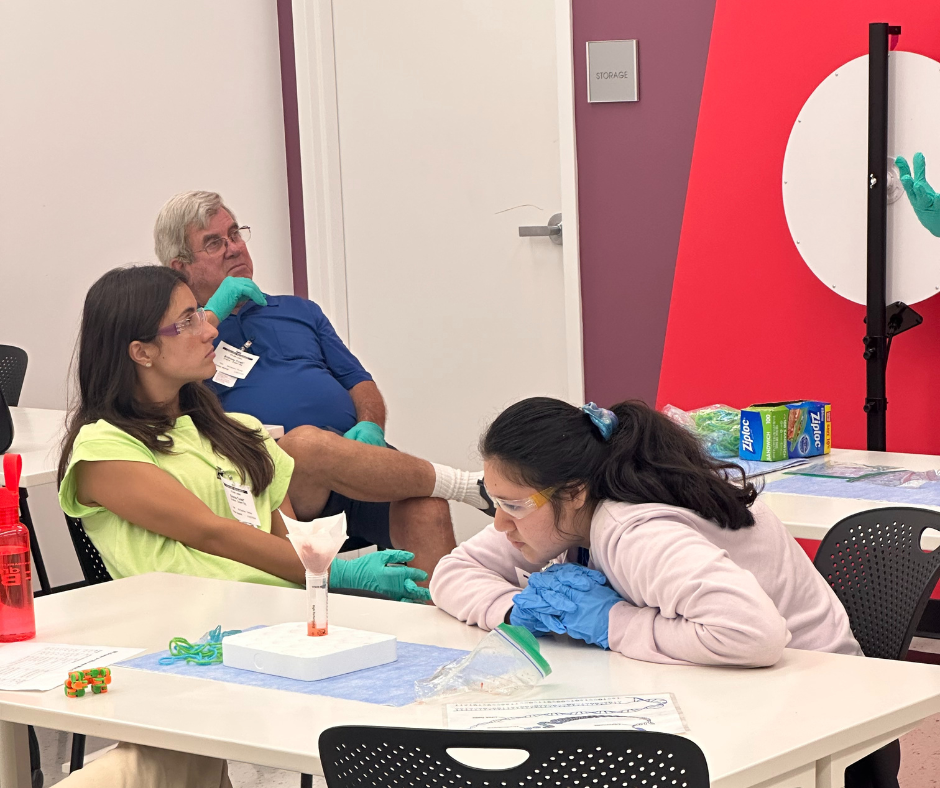 Emma and Tony at Bayer In a science learning lab, a student observes an experiment as Camp counselor and driver listen to an instructor.