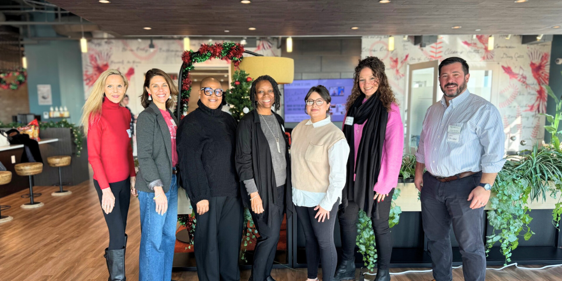 Seven professionals smile together in an office community room.