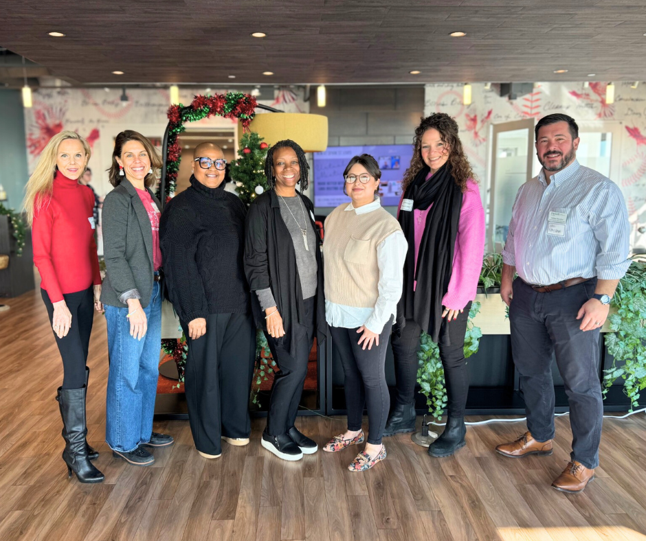 Seven professionals smile together in an office community room.